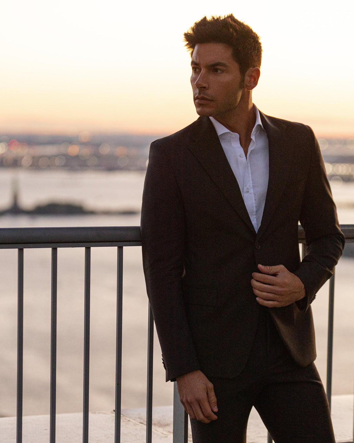 A man in a black suit and open-collar white shirt at sunset