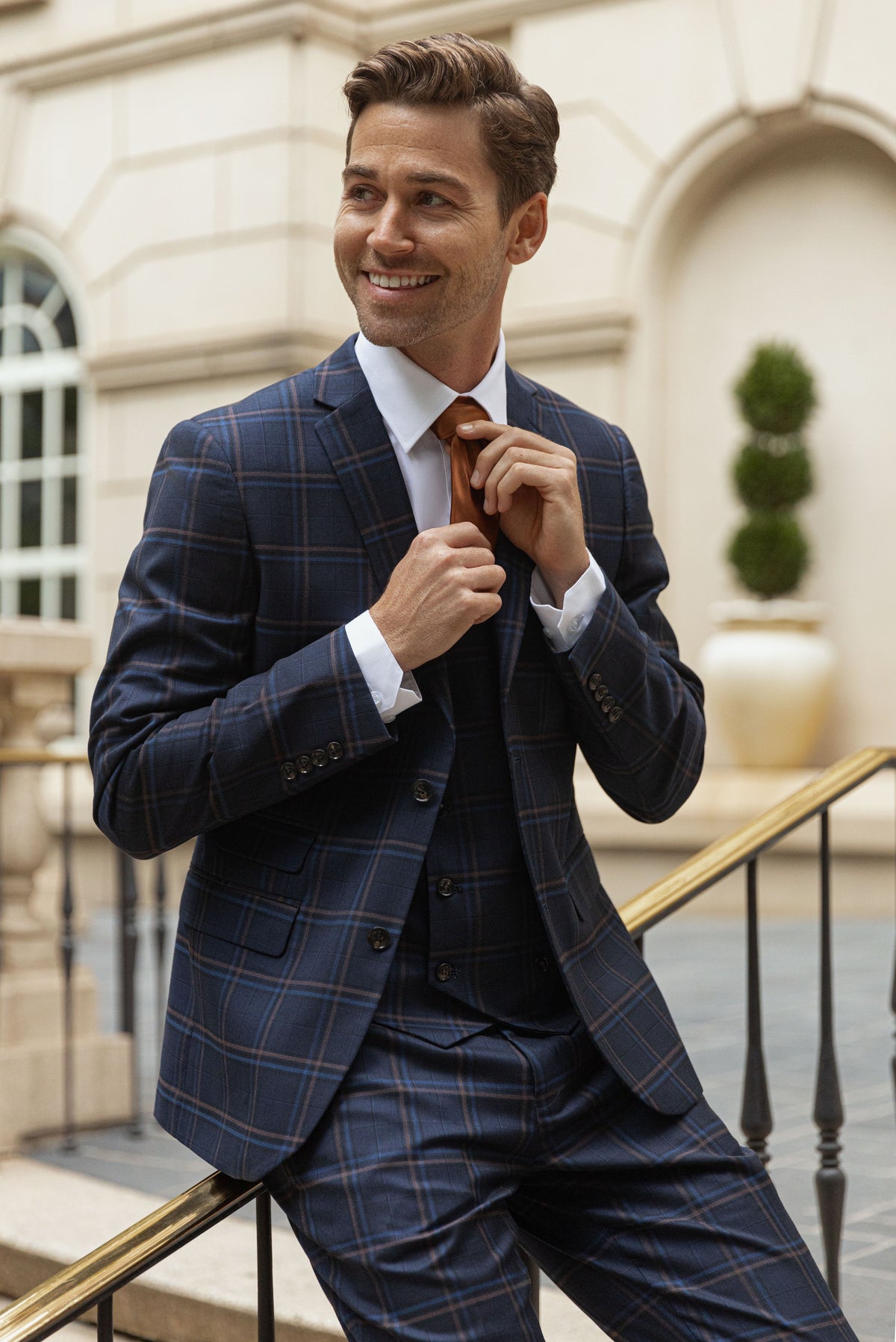A smiling man adjusting his red tie in a navy three-piece suit on classic stone steps