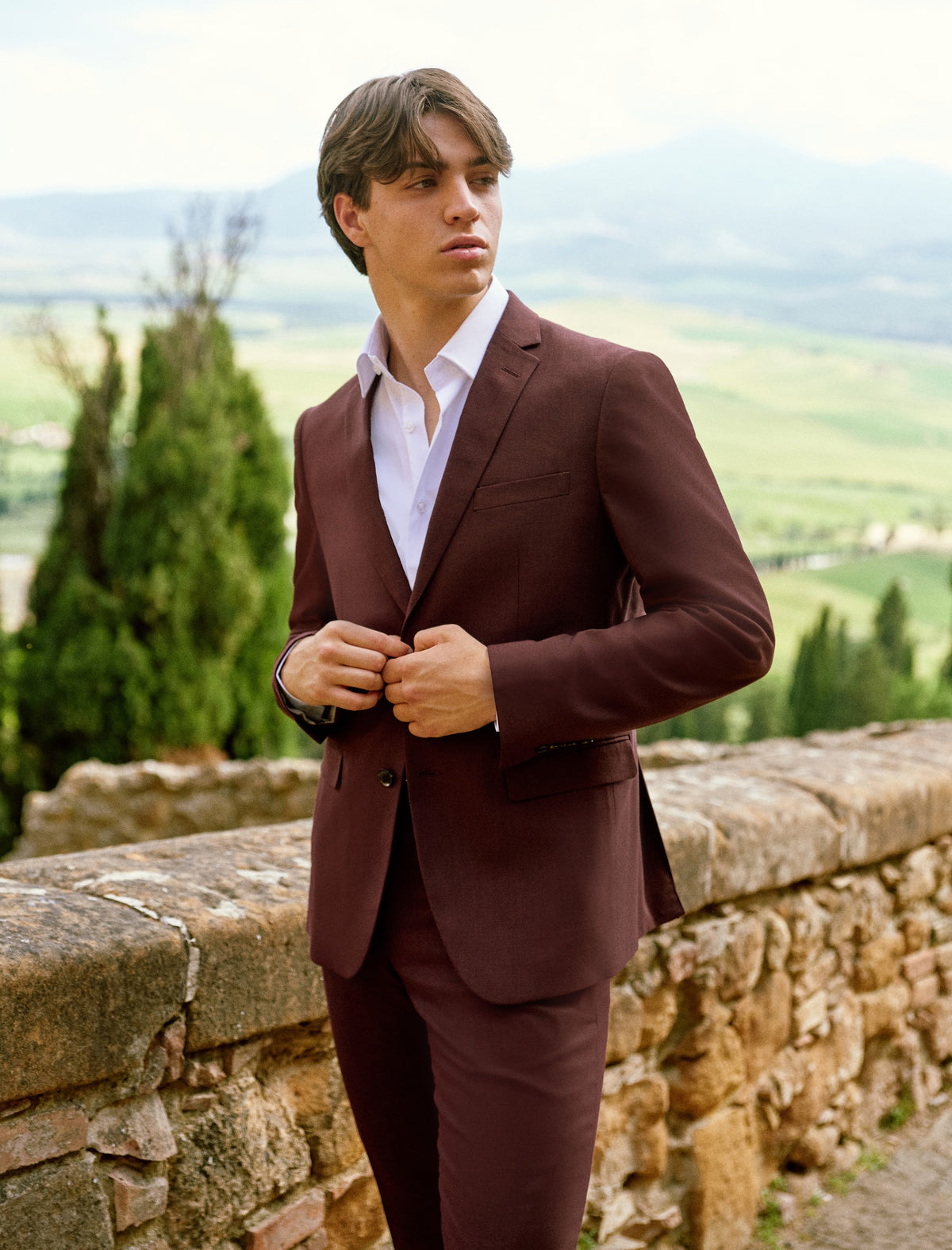 A young man in a burgundy suit on a stone wall in Tuscany