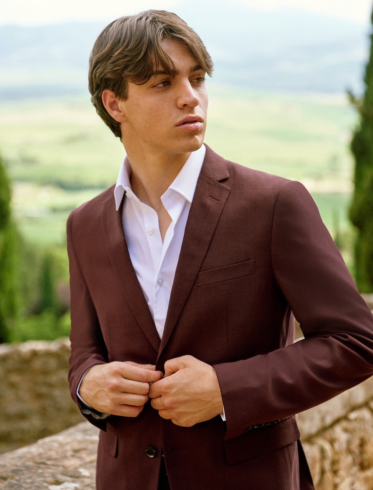 A young man in a burgundy suit with the Tuscan hills behind