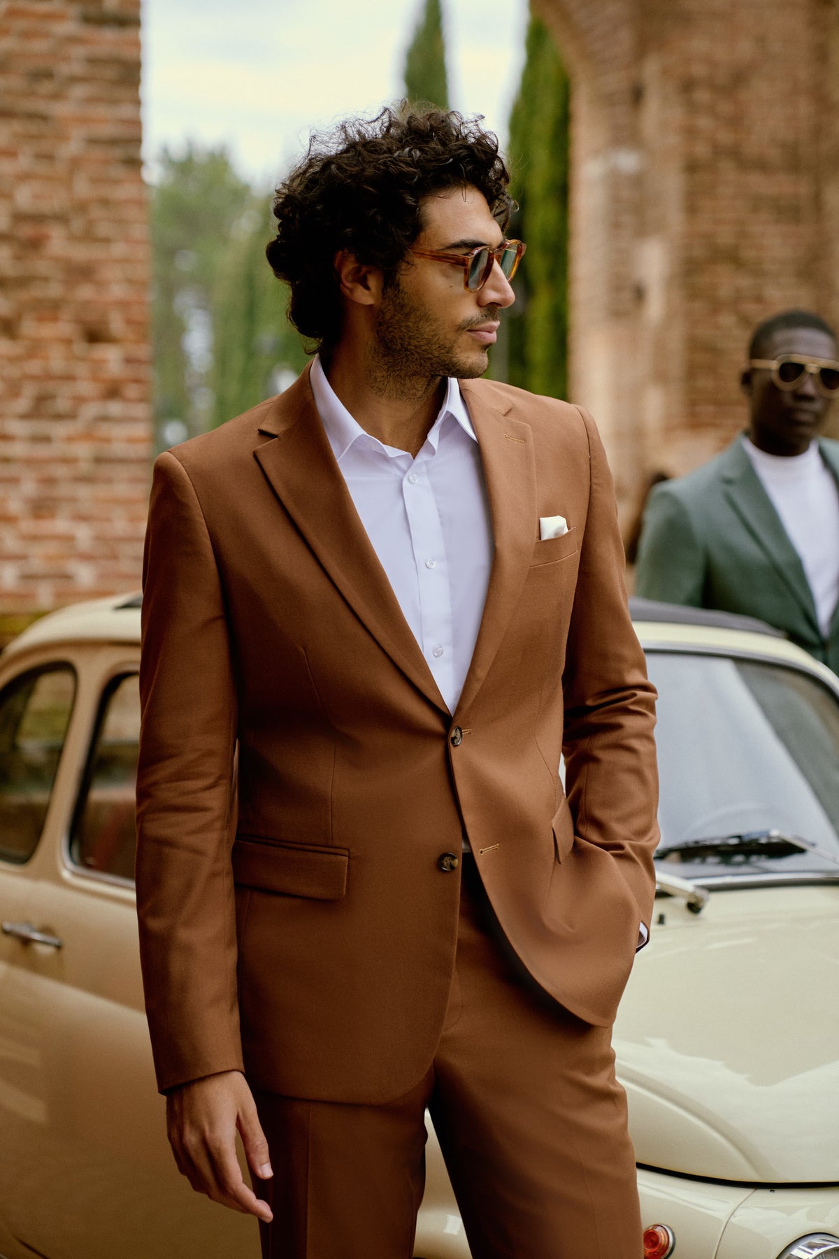 A man in a cognac suit beside a vintage car in Italy