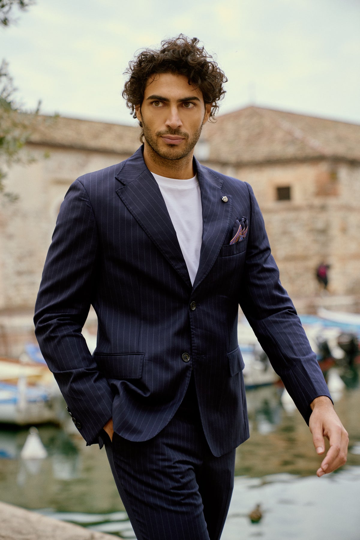 A man in a navy suit and white tee by a harbor in Italy