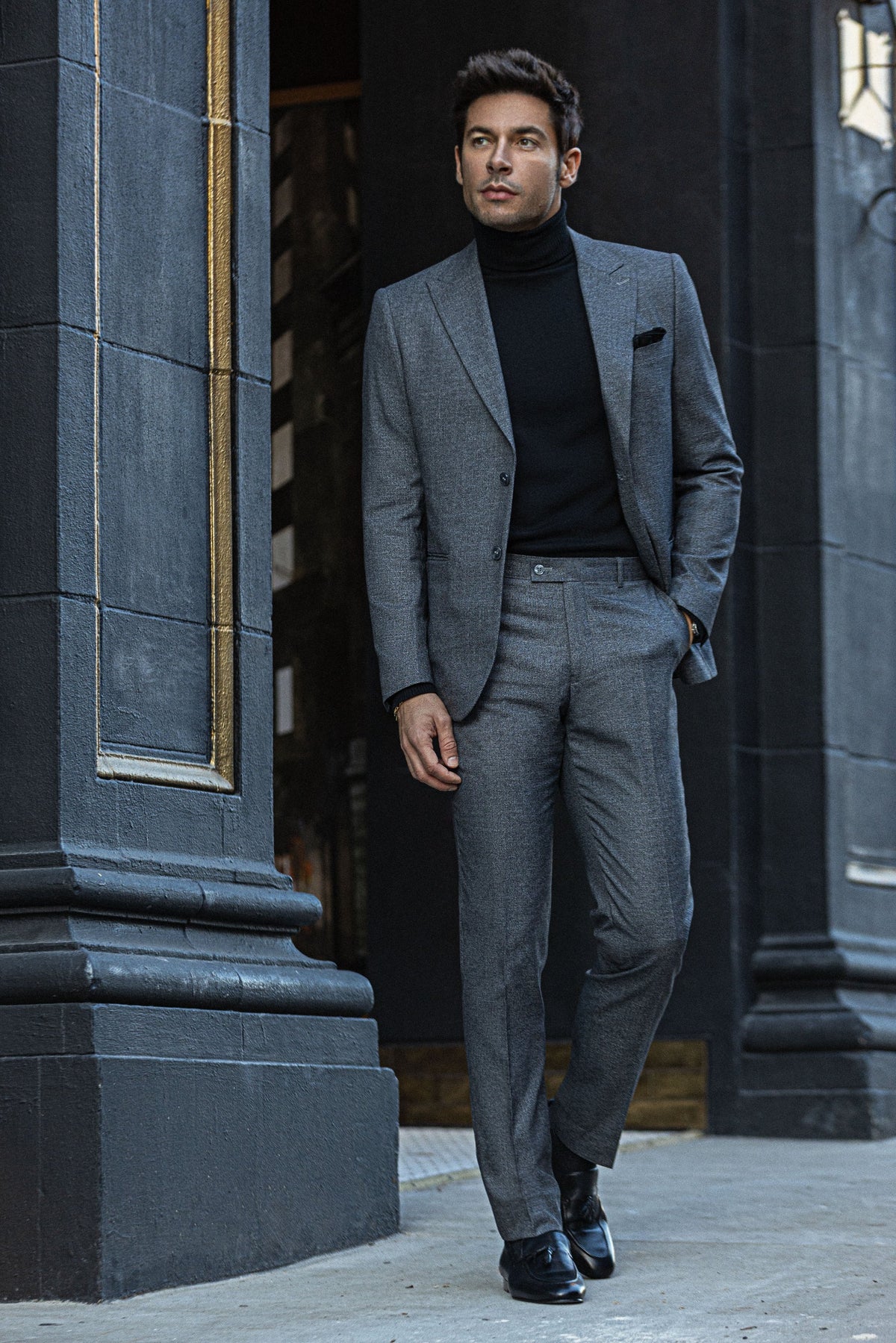 A man in a gray suit and black turtleneck between stone columns
