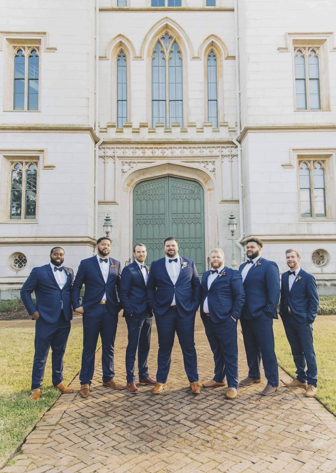 Seven groomsmen in navy suits and bow ties in front of a historic stone building